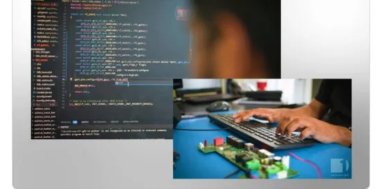 A monitor showing embedded C firmware open in an editor, with a photograph inset over one corner of an engineer's hands at a keyboard beside a populated development board on a blue mat.