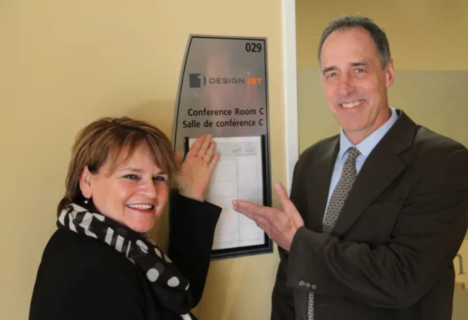 Joan Highet and Kevin Bailey beside the Design 1st conference room sign at United Way Ottawa