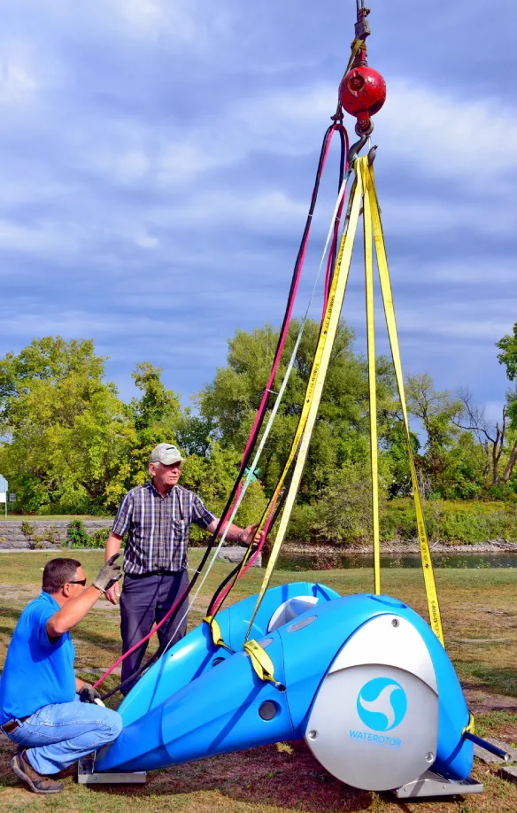 Two technicians rigging a blue Waterotor turbine unit to a crane sling beside a river