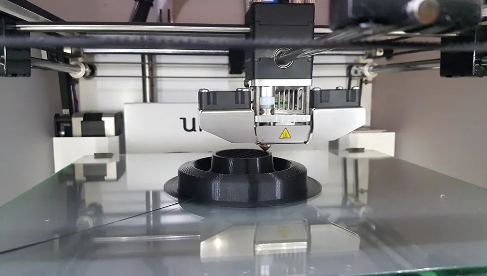 Ultimaker 3D printer extruding a black coffee filter holder on its glass bed