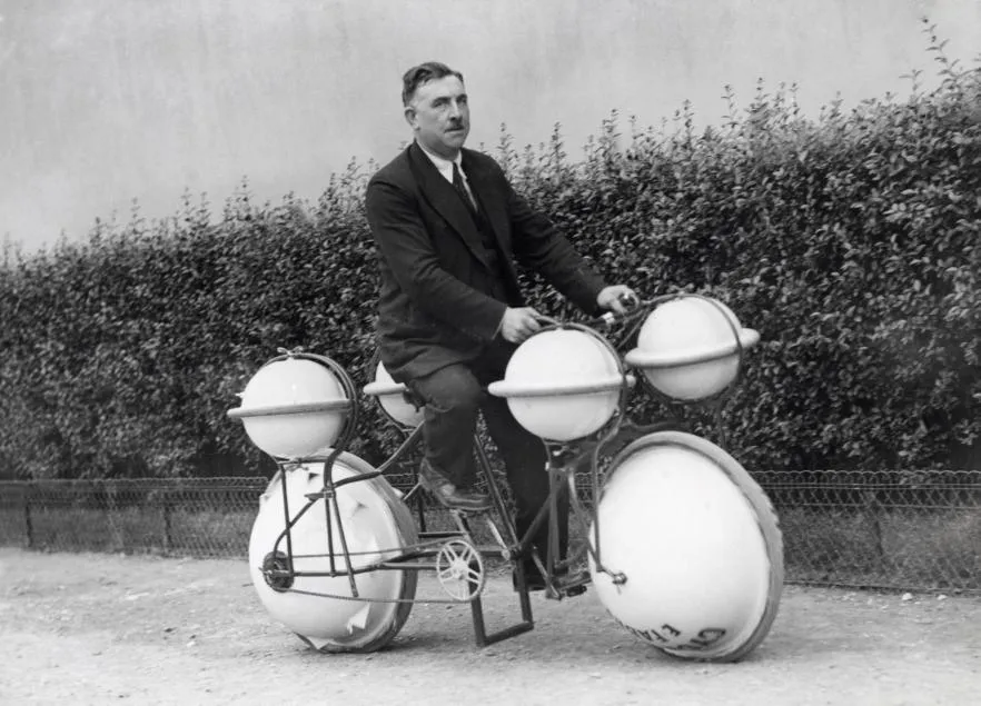 Black-and-white photo of a man riding an amphibious bicycle fitted with four large floats