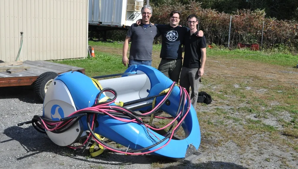 Three Design 1st mechanical engineers behind a blue Waterotor hydrokinetic turbine prototype outdoors
