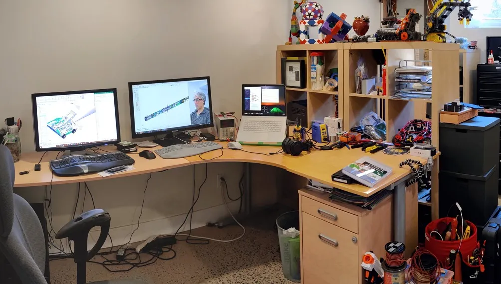 Home office workstation with three monitors showing CAD models, test equipment and components on the desk