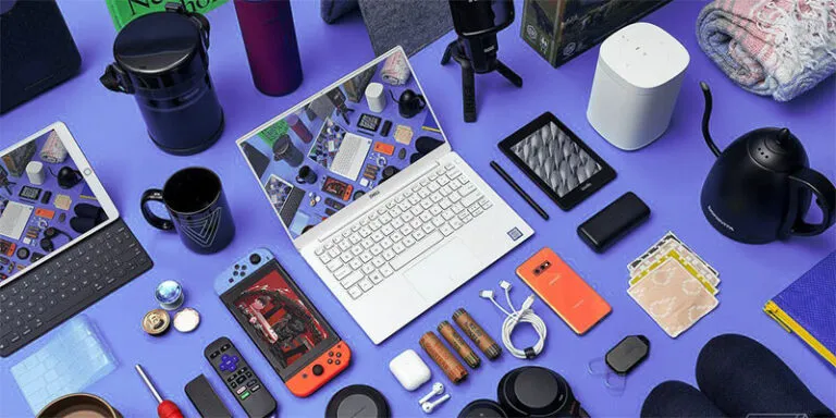 Flat lay of consumer gadgets including a laptop, tablet, game console, earbuds and kettle on a purple background