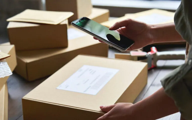 Online seller scanning a labelled shipping box with a smartphone beside stacked parcels