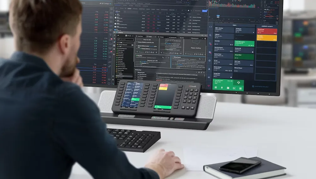 Trader at a desk using the Cloud9 VoIP device beneath a large trading screen