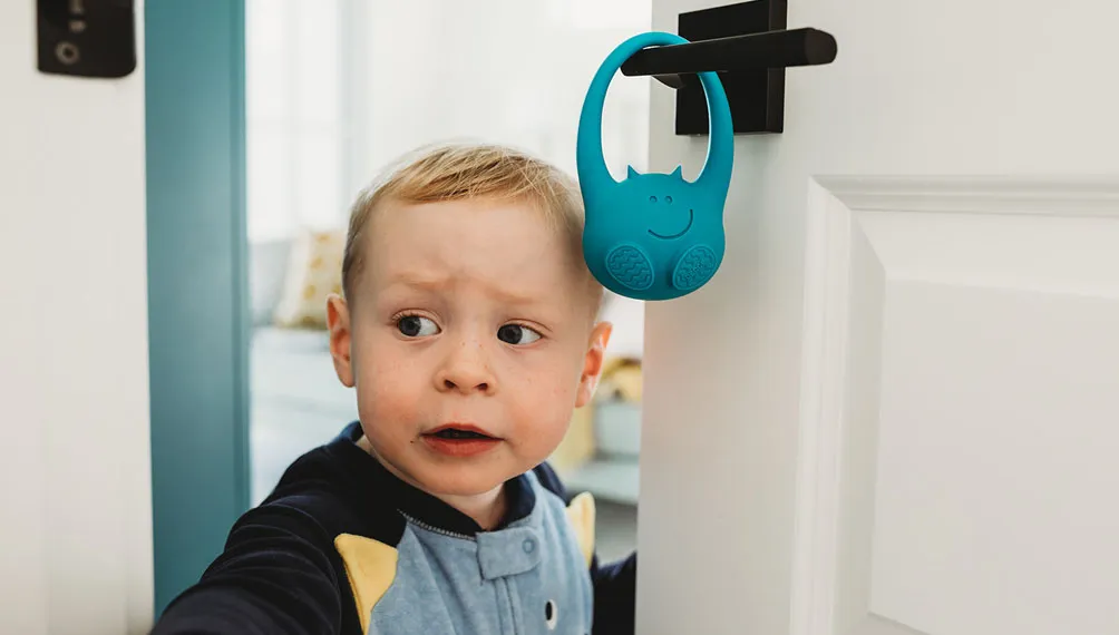 Toddler peeking around a door with the blue toddlermonitor hanging on the handle