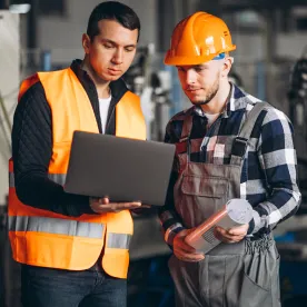 Two engineers in safety vests reviewing a laptop and a sample part on a factory floor